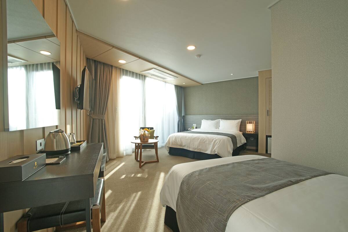Habitación de hotel con una cama grande, una cama más pequeña, un escritorio con una silla, una mesa con una lámpara y un espejo grande.