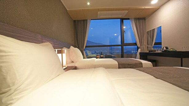Habitación de hotel con dos camas, un escritorio y un ventanal con vista a la ciudad al atardecer.