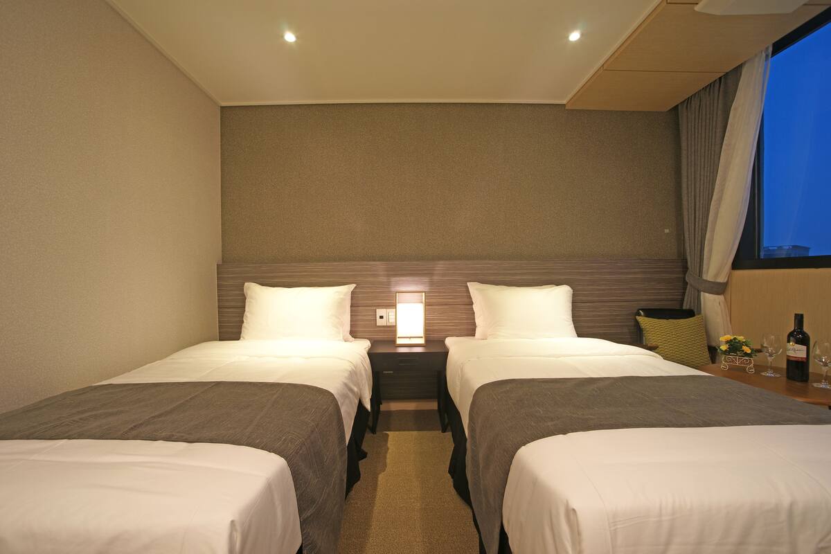 Una habitación de hotel con dos camas individuales, una mesita de noche con lámpara, una pequeña mesa con una botella y vasos, y una ventana con cortinas.