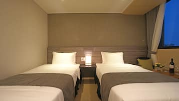 Ein Hotelzimmer mit zwei Einzelbetten, einem Nachttisch mit Lampe, einem kleinen Tisch mit Flasche und Gläsern sowie einem Fenster mit Vorhängen.