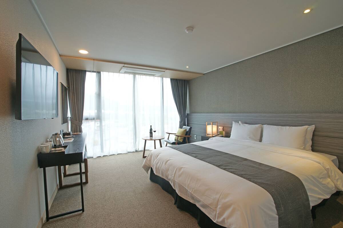 Habitación de hotel con una cama grande, un escritorio con silla, una mesita con una botella y un televisor en la pared.