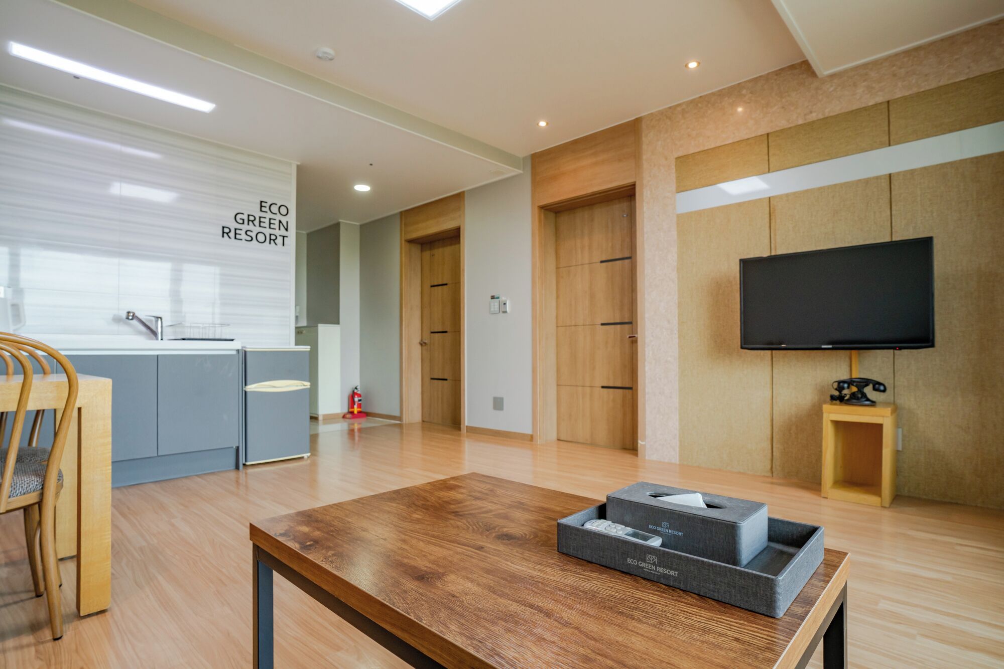 A modern living room with a wooden coffee table, a flat-screen TV, and a kitchen area with a sink and cabinets.