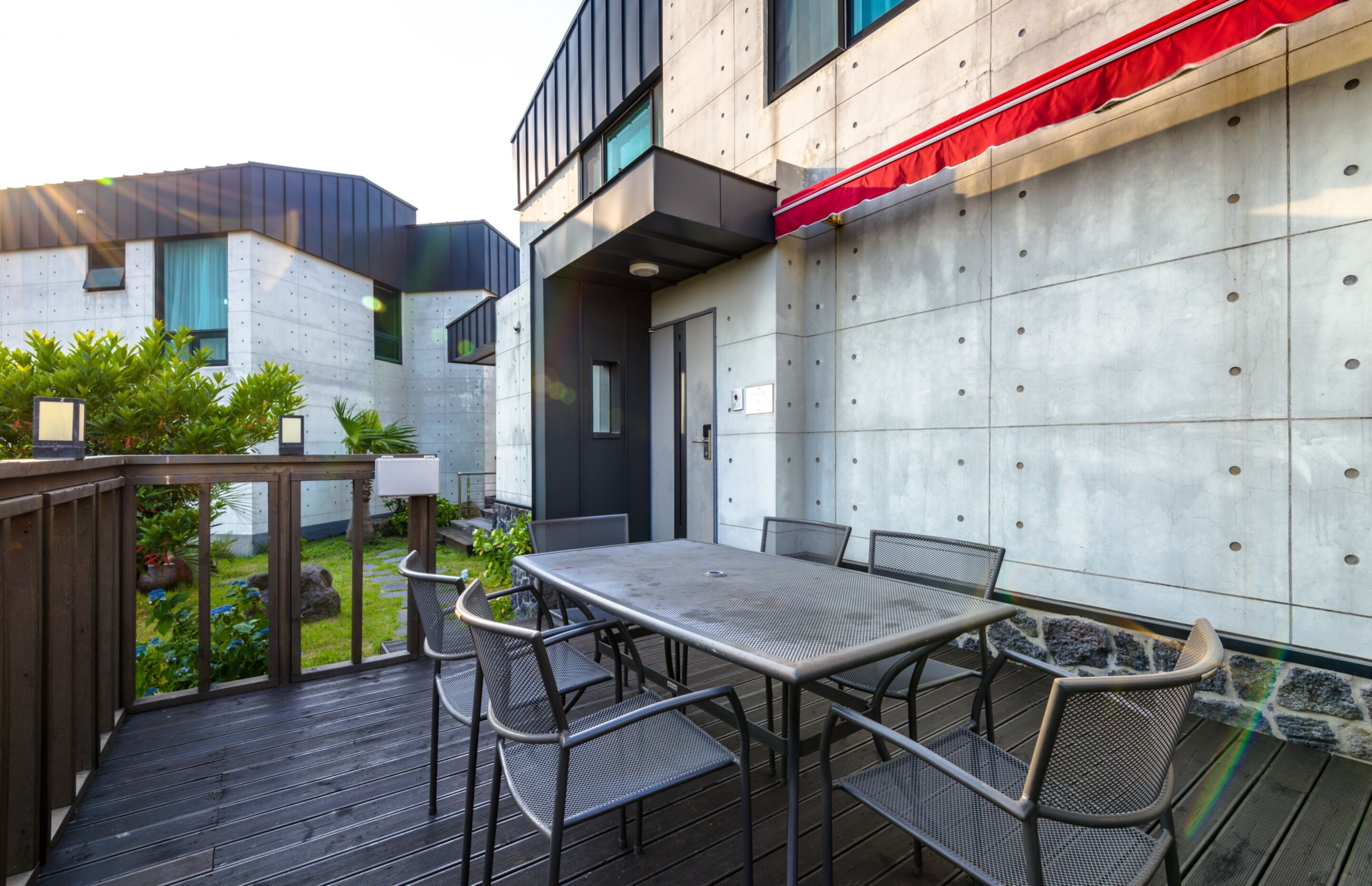 Um pátio externo com mesa e cadeiras de metal, um deck de madeira e vista para um edifício moderno com toldo vermelho.