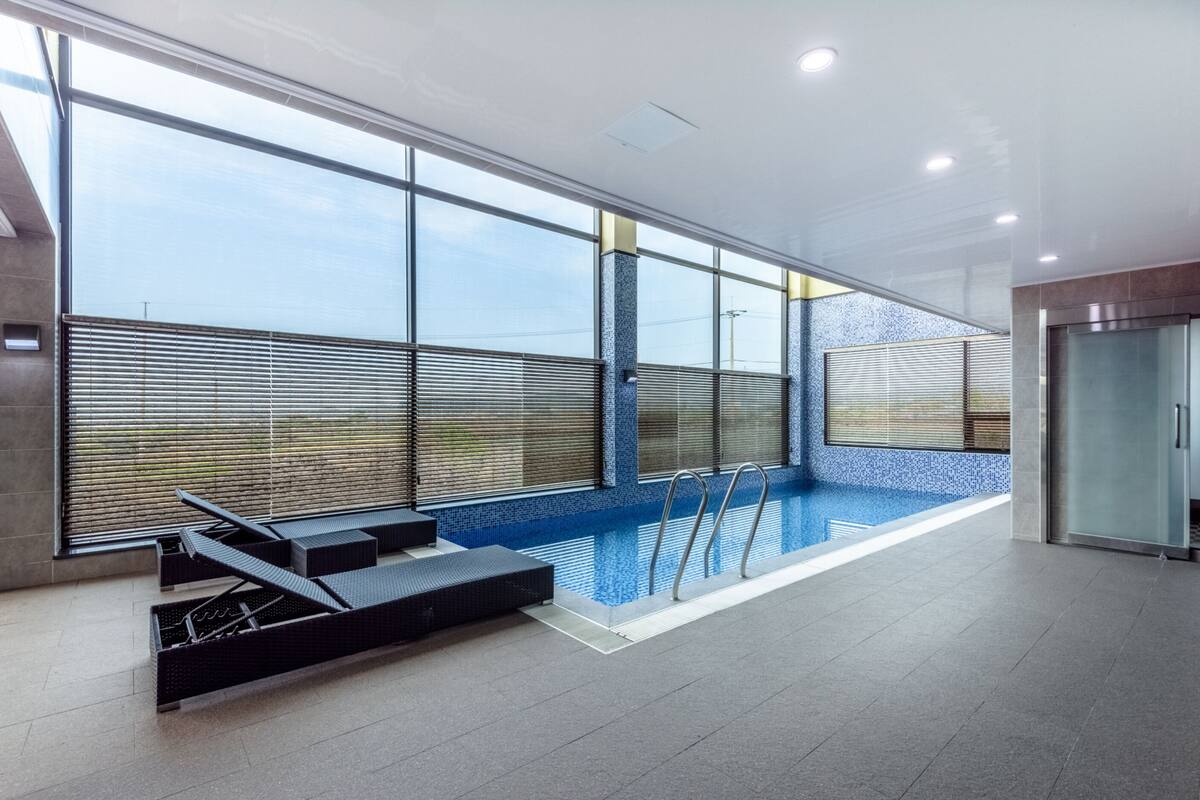 A modern indoor swimming pool with clear glass walls, a tiled floor, and a glass elevator.