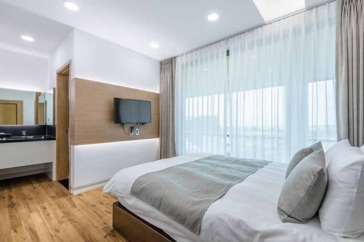 A modern hotel room with a large bed, a flat-screen TV mounted on the wall, and a large window with sheer curtains.