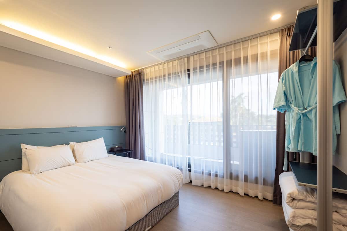 A hotel room with a large bed, a bedside table, a robe hanging on a rack, and a view through sheer curtains.
