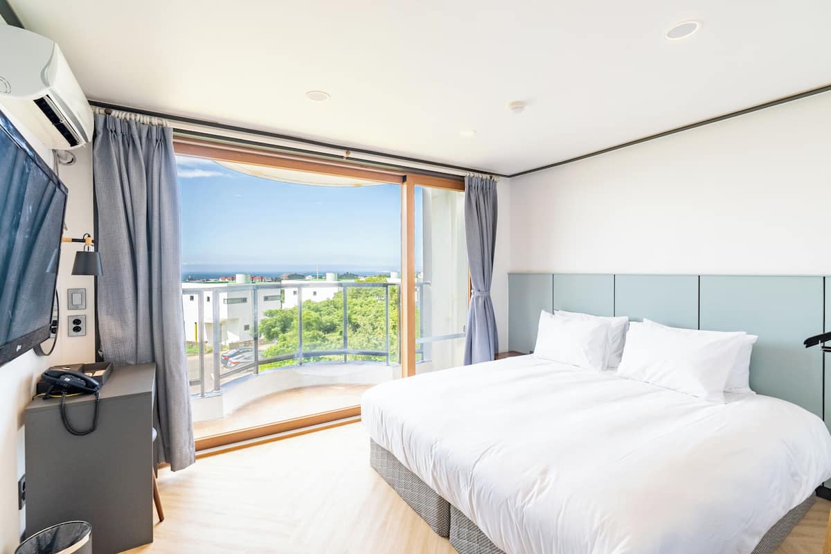 A hotel room with a large bed, a desk with a phone, and a view of the city and sea through a balcony.