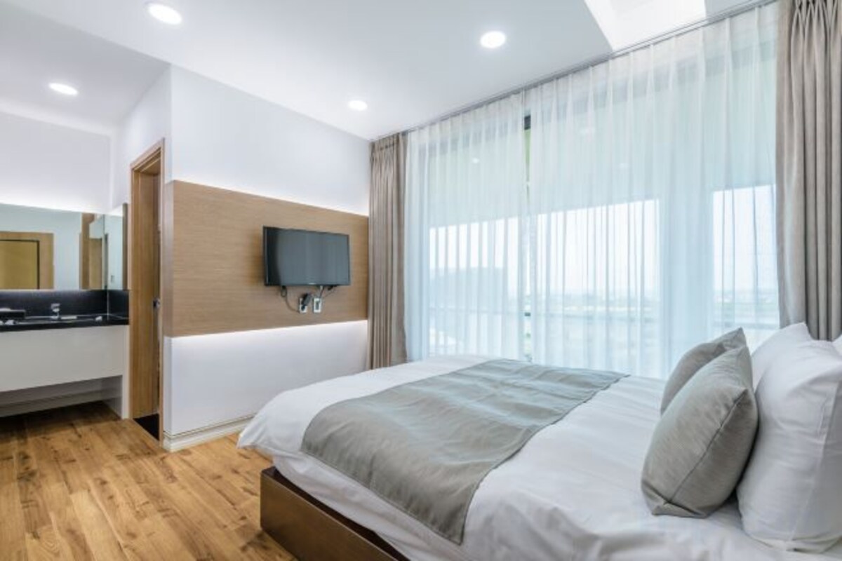 Une chambre d’hôtel moderne avec un grand lit, un téléviseur à écran plat fixé au mur et une grande fenêtre agrémentée de rideaux légers.