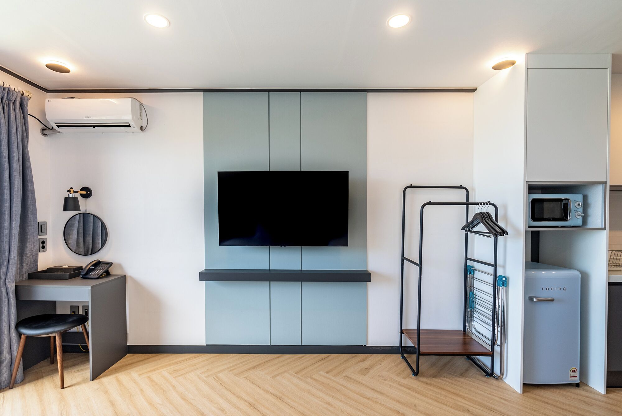 Una sala de estar moderna con un televisor de pantalla plana montado en la pared, un escritorio pequeño con una silla, un perchero y una kitchenette compacta con microondas.