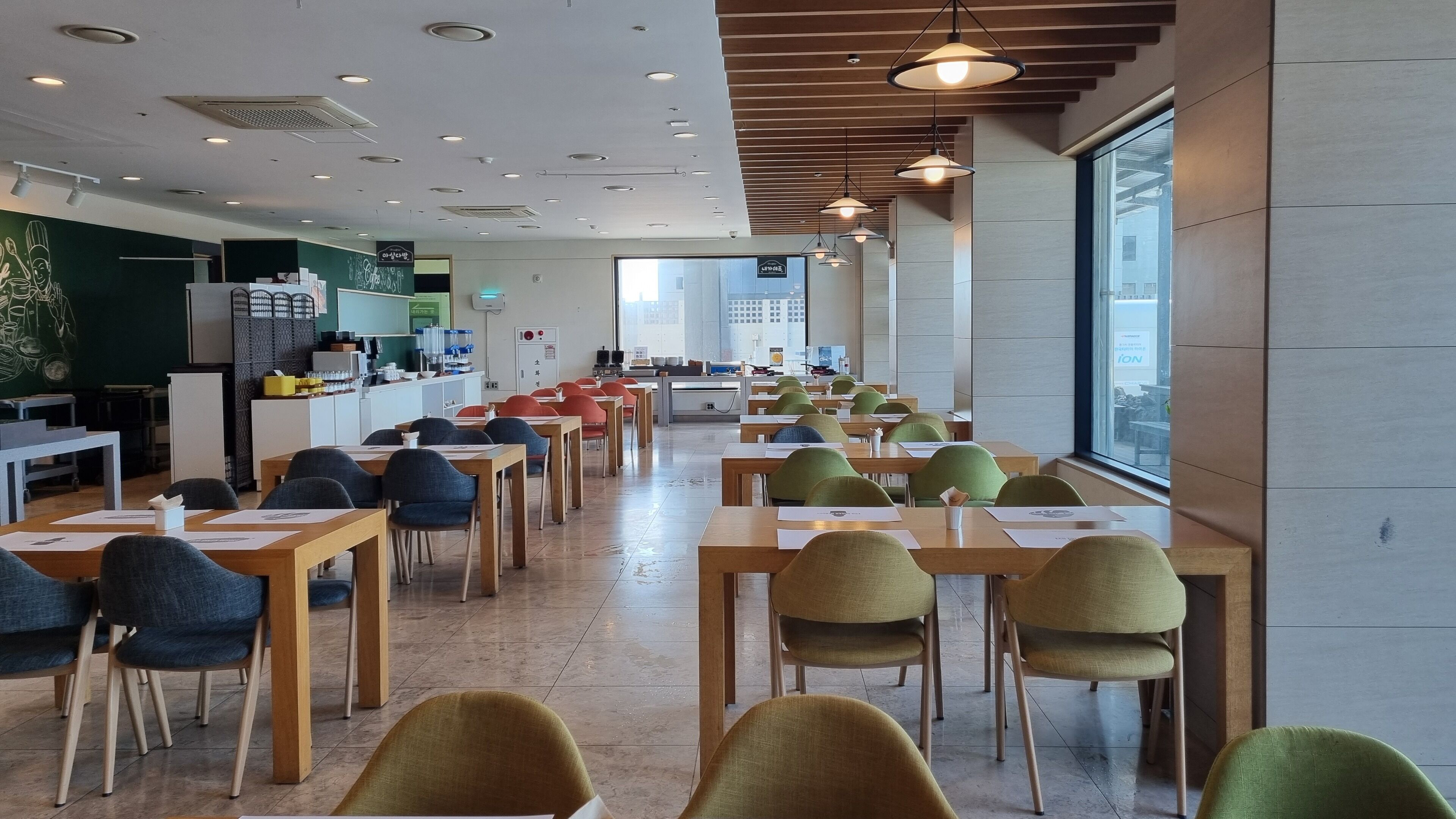 A spacious dining area with long tables and chairs, a kitchen area with a chalkboard menu, and large windows providing a view of the city.