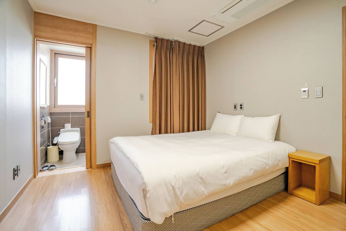 A bedroom with a bed, a bedside table, a window with curtains, a bathroom with a toilet, and a shower area.