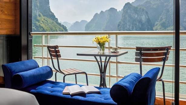 Ein Balkon mit Blick auf eine zerklüftete Küstenlandschaft, einem Tisch mit Blumenvase und blau gepolsterten Stühlen.