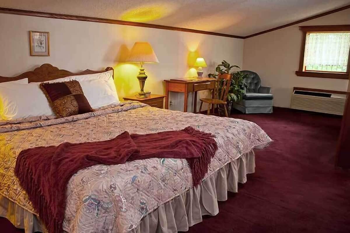A neatly made bed with a patterned bedspread and pillows, a bedside table with lamps, a chair, and a radiator.