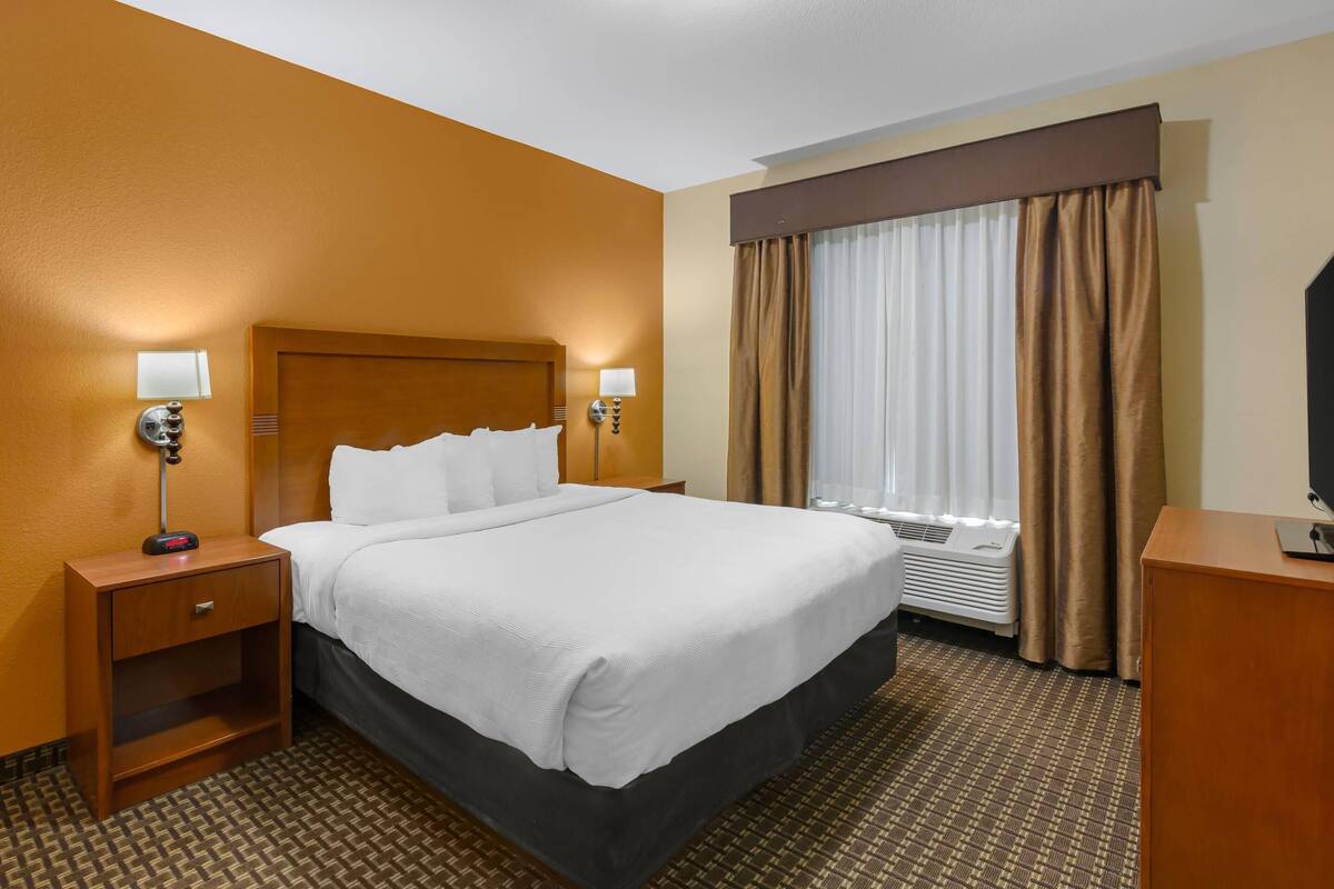A hotel room with a large bed, a nightstand with a lamp, a desk with a computer, and a window with curtains.