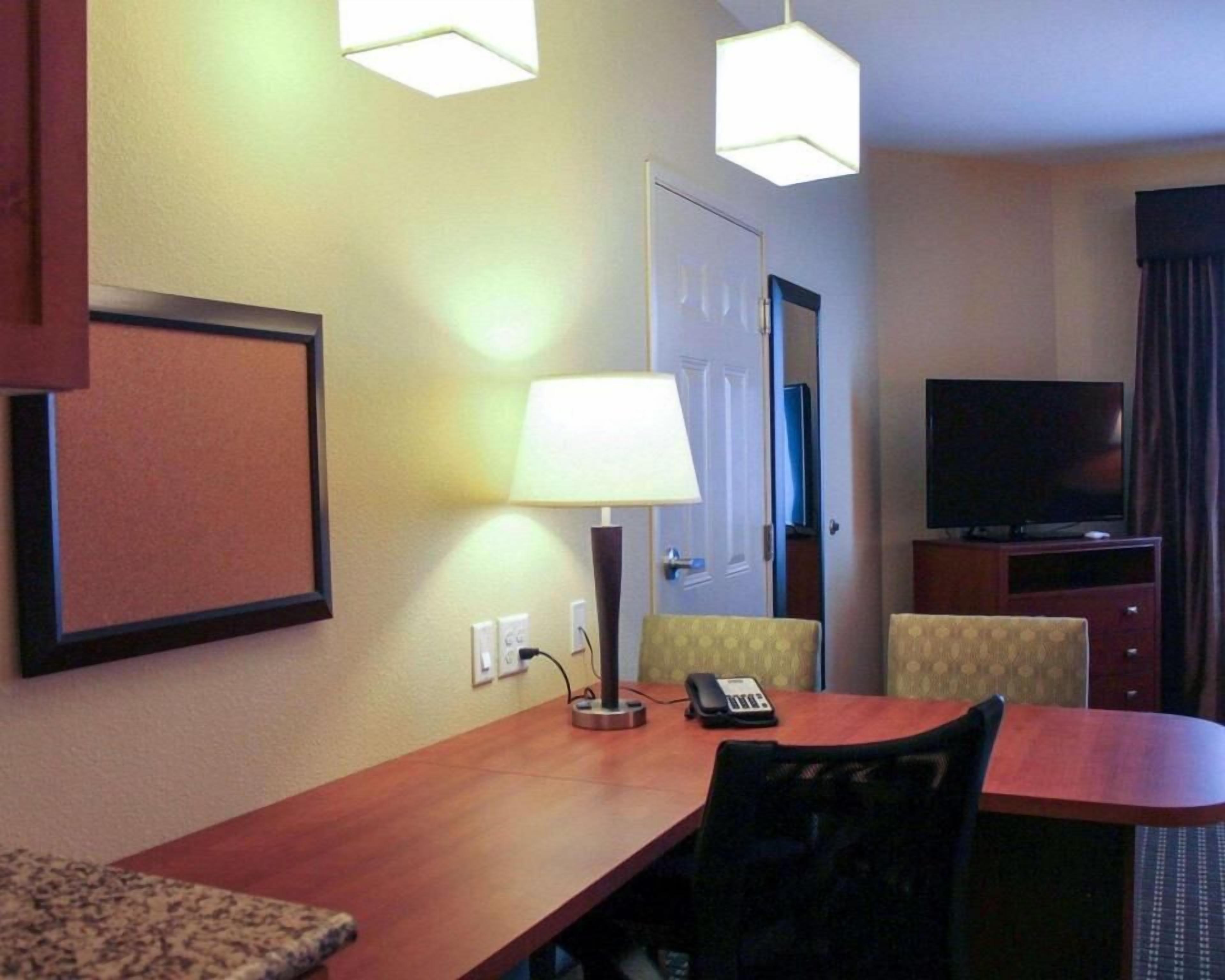 A hotel room with a wooden desk, a chair, a lamp, a telephone, a television, and a corkboard on the wall.