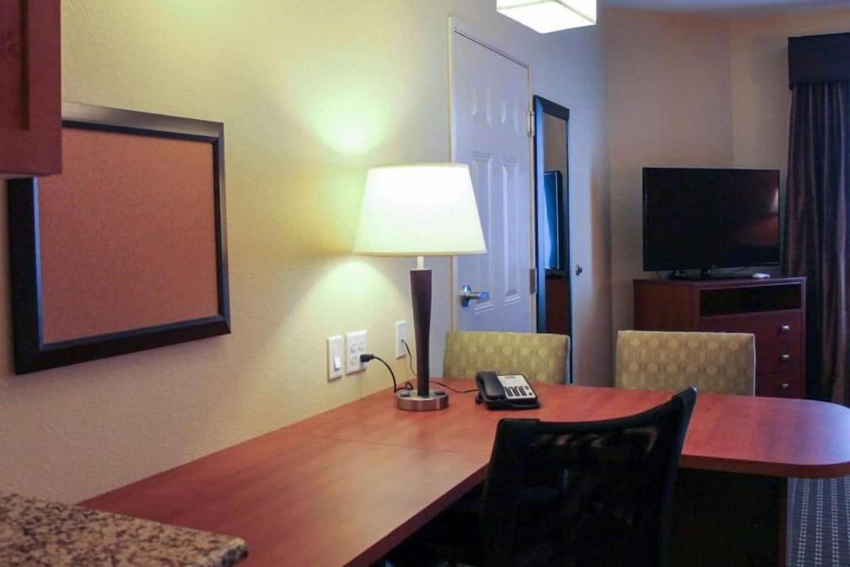 A hotel room with a wooden desk, a chair, a lamp, a telephone, a television, and a corkboard on the wall.