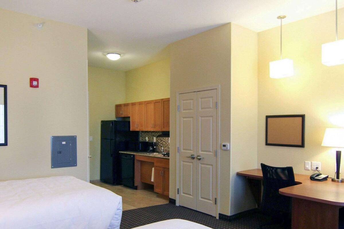 A hotel room with two beds, a kitchenette, a desk with a chair, and a small table with a lamp.