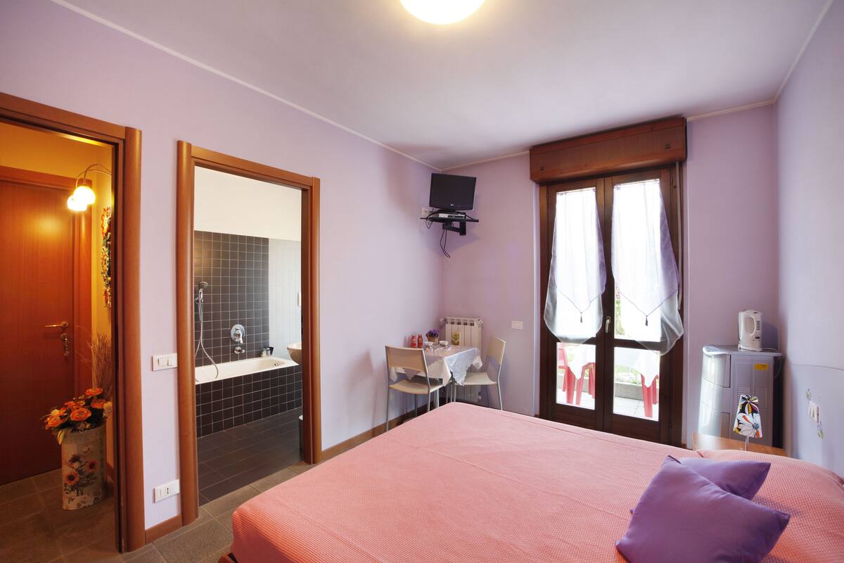 A bedroom with a bed, a television mounted on the wall, a window with curtains, and a bathroom visible through an open door.