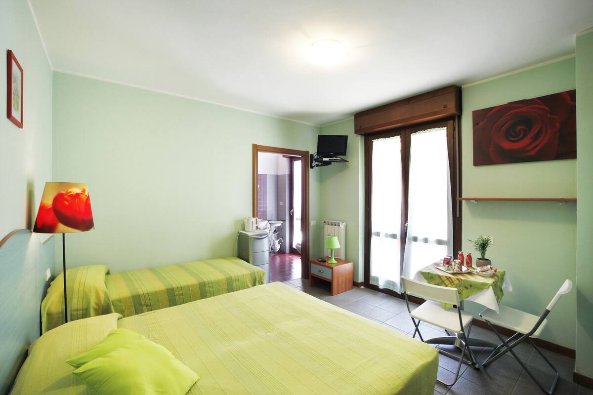 A room with two beds, a small table with a tablecloth, a chair, a television on the wall, and a door leading to another room.