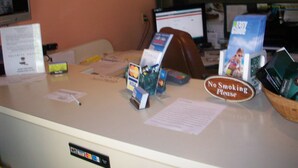 A reception desk with a 'No Smoking' sign, a computer monitor, and various informational brochures. - All Suite Motel (Edgewater)