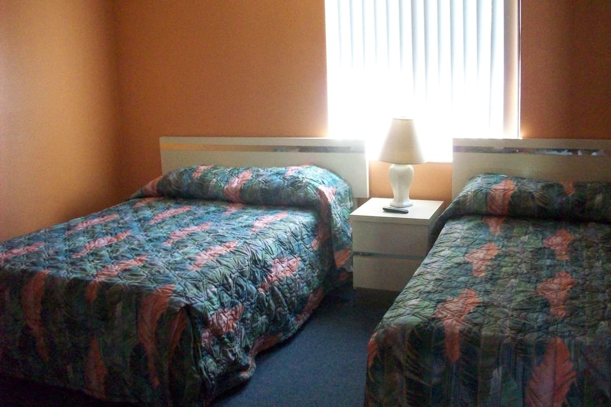 Una habitación con dos camas, cada una con una colcha de motivos florales, una mesita de noche con lámpara y una ventana con persianas.