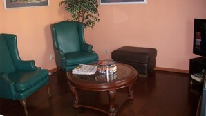 A living room with a green armchair, a brown ottoman, a round wooden coffee table, and a potted plant. - All Suite Motel (Edgewater)