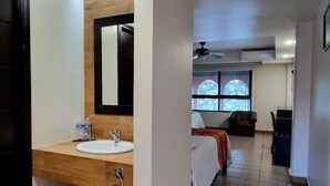 Quarto de hotel com piso de madeira, um espelho grande sobre a pia, uma cama com colcha branca e uma janela com vista para árvores.