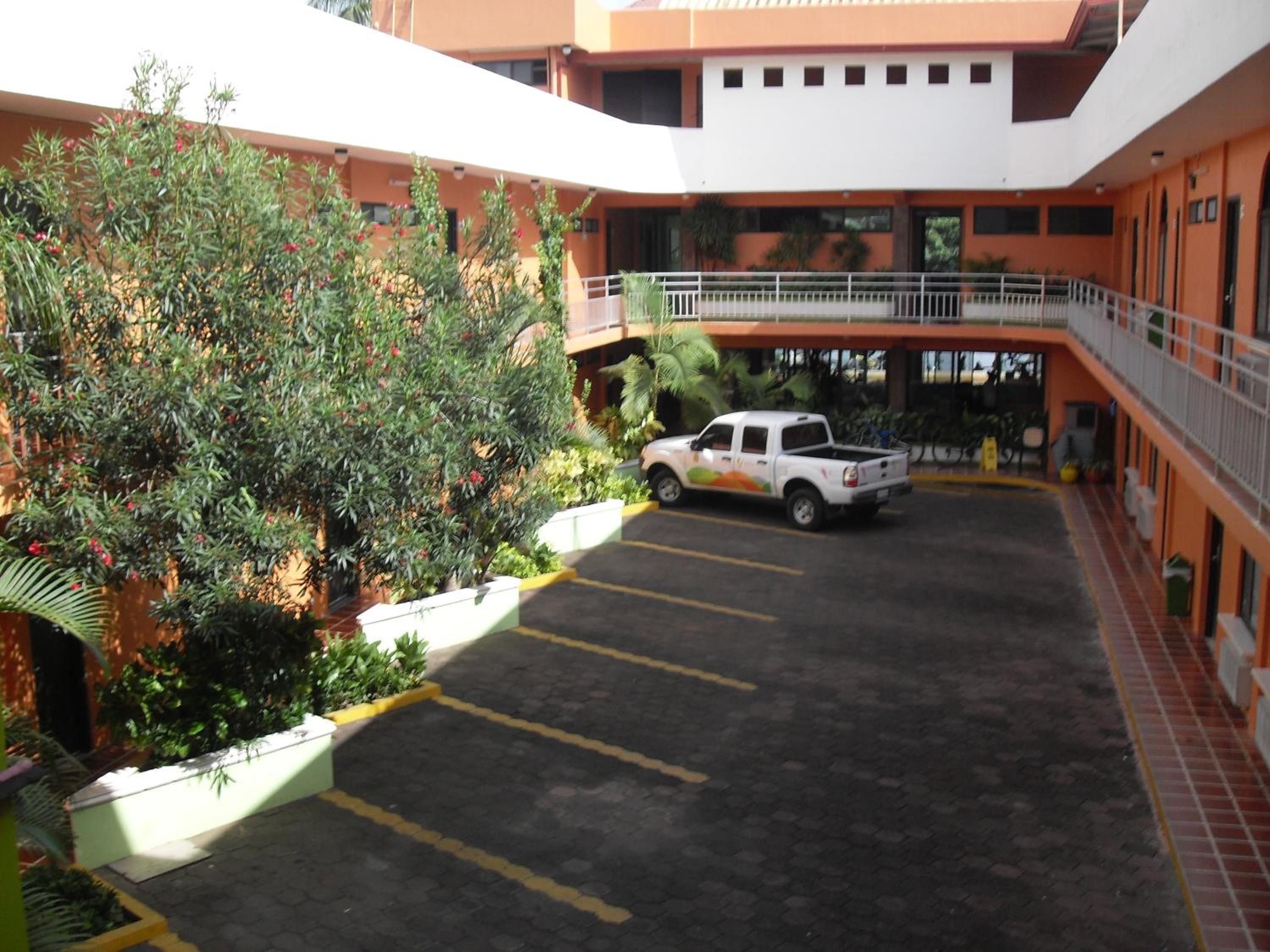 A multi-level parking area with a white pickup truck parked in the center, surrounded by greenery and a building with orange and white walls.