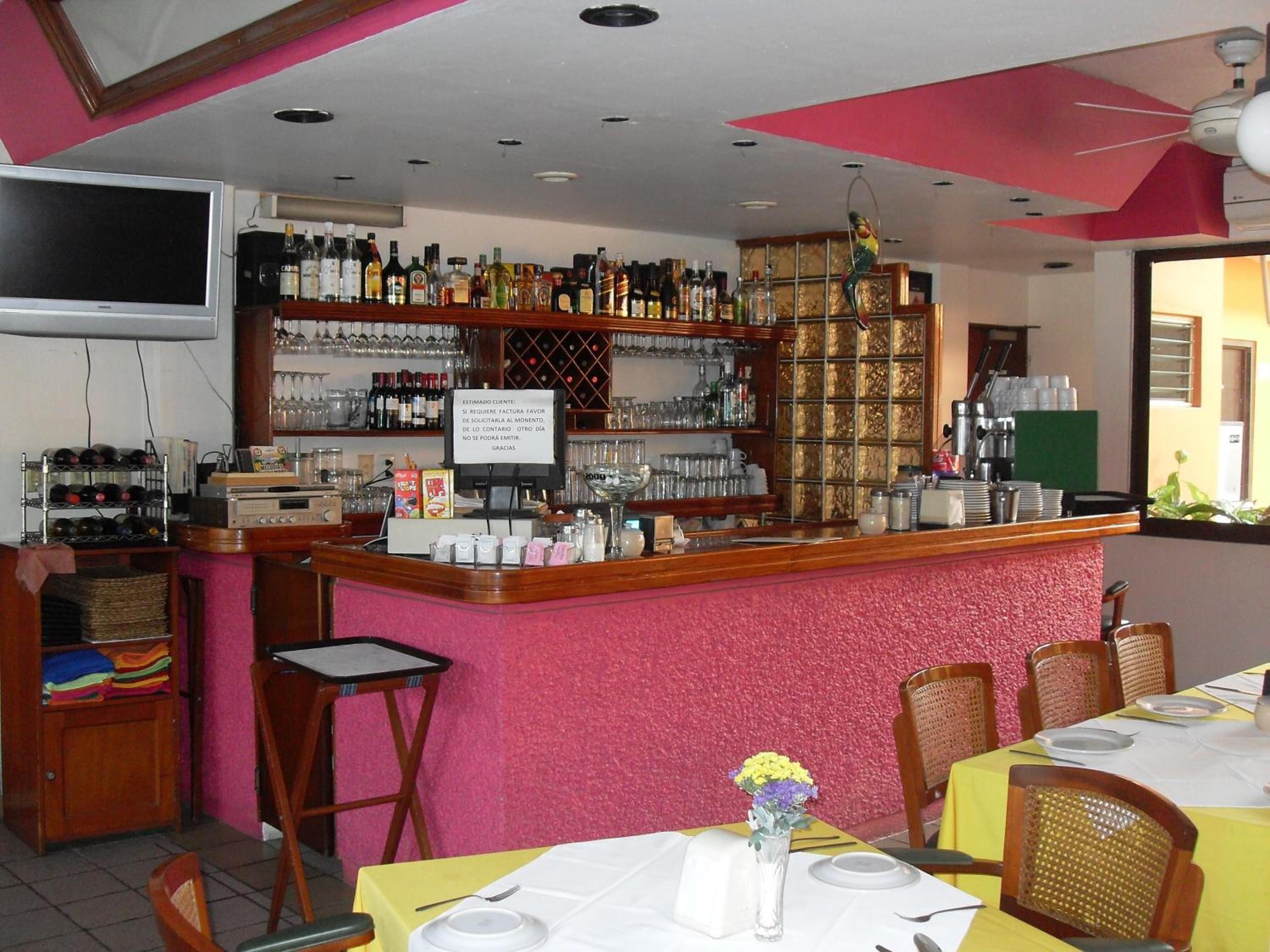 Angolo bar con scaffali pieni di bottiglie e bicchieri, una televisione e un cartello in spagnolo.
