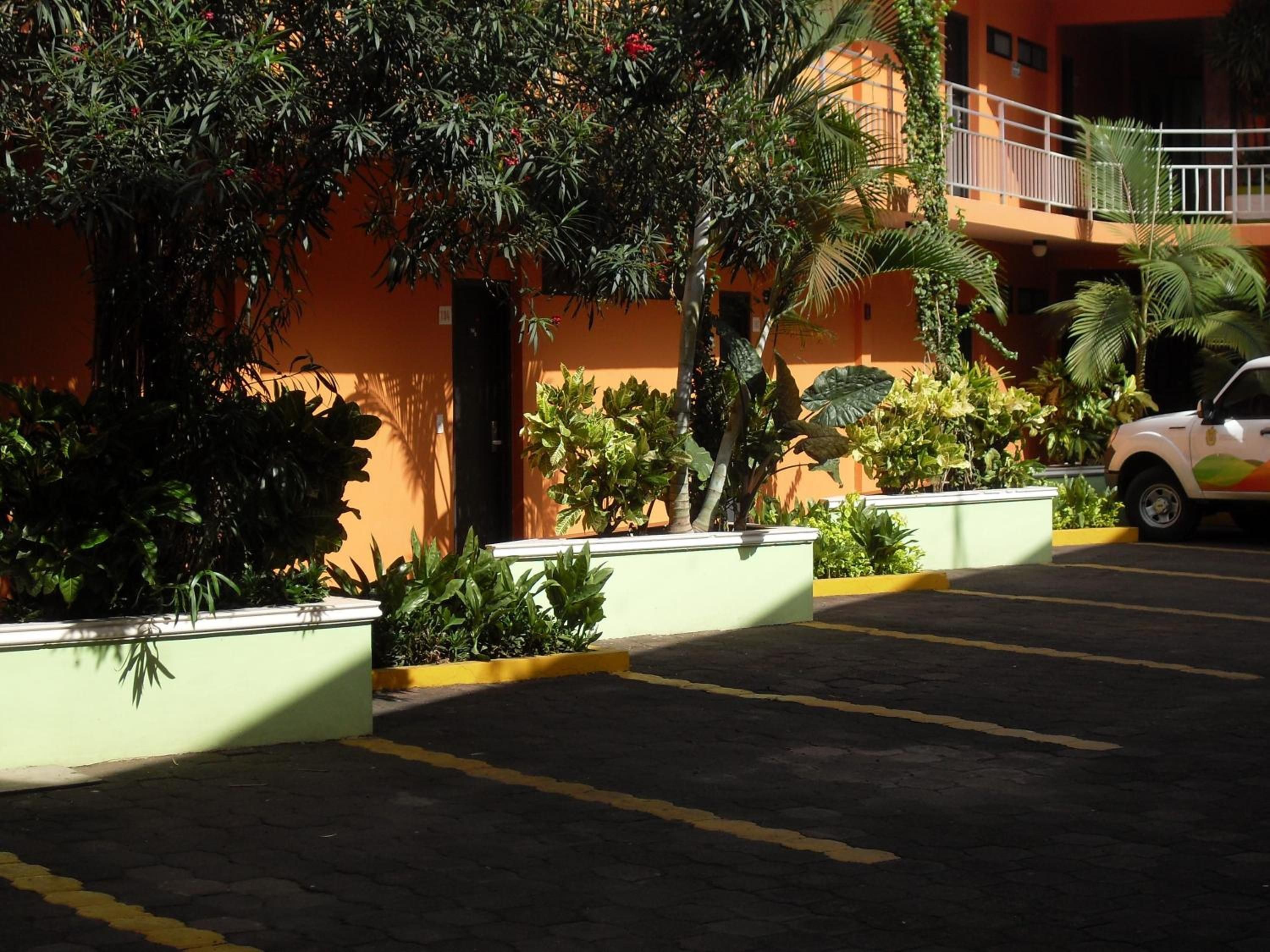 Un estacionamiento con un coche blanco, rodeado de vegetación y un edificio de fachada naranja.
