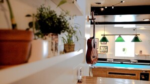 Una mesa de billar con mesa y sillas de madera, rodeada de plantas en macetas y lámparas verdes colgantes.