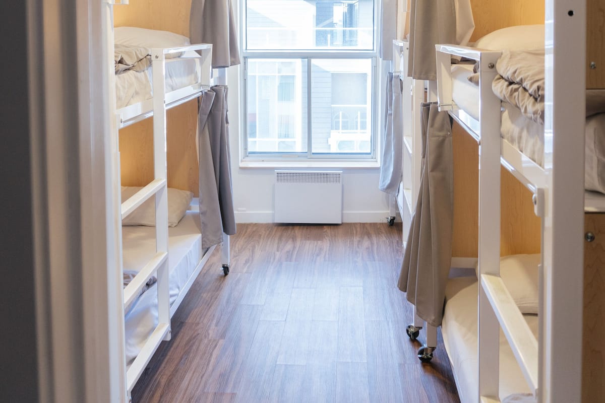 A narrow corridor with bunk beds on either side, a window with curtains, and a wooden floor.