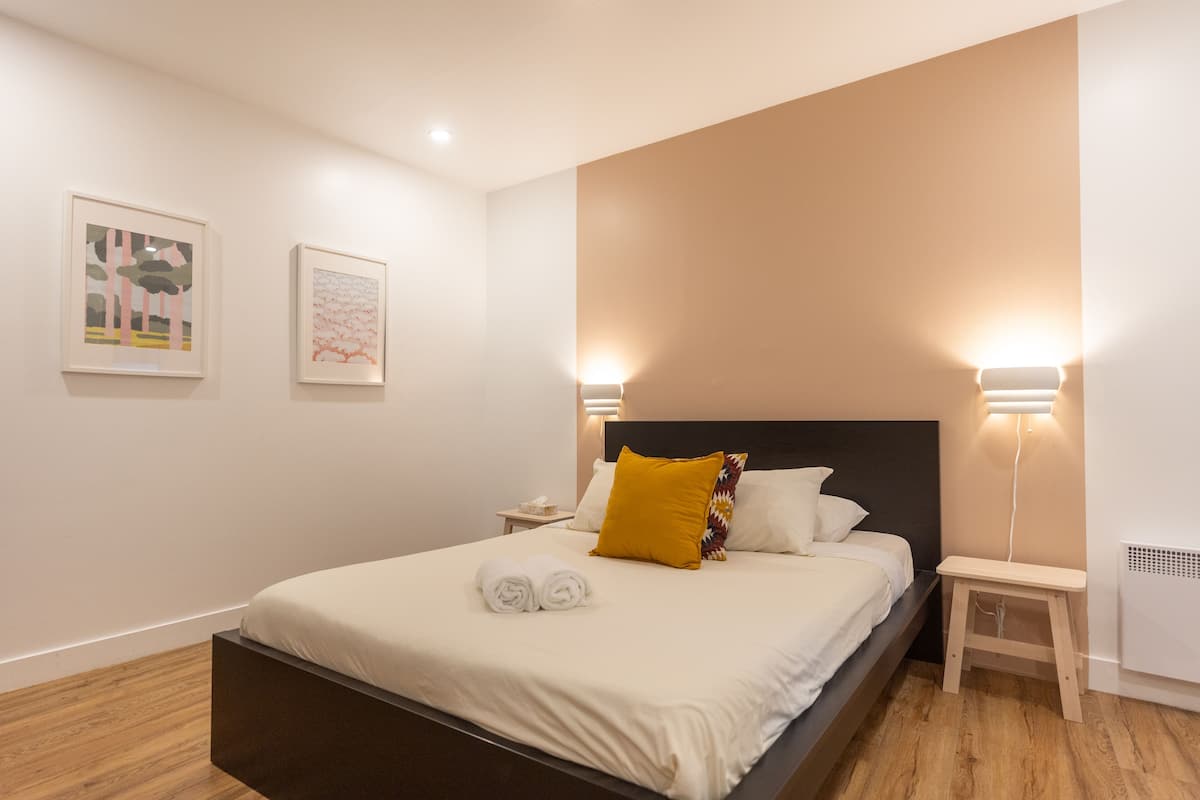 A neatly made bed with white linens and a rolled towel, a bedside table with a lamp, and two framed artworks on the wall.