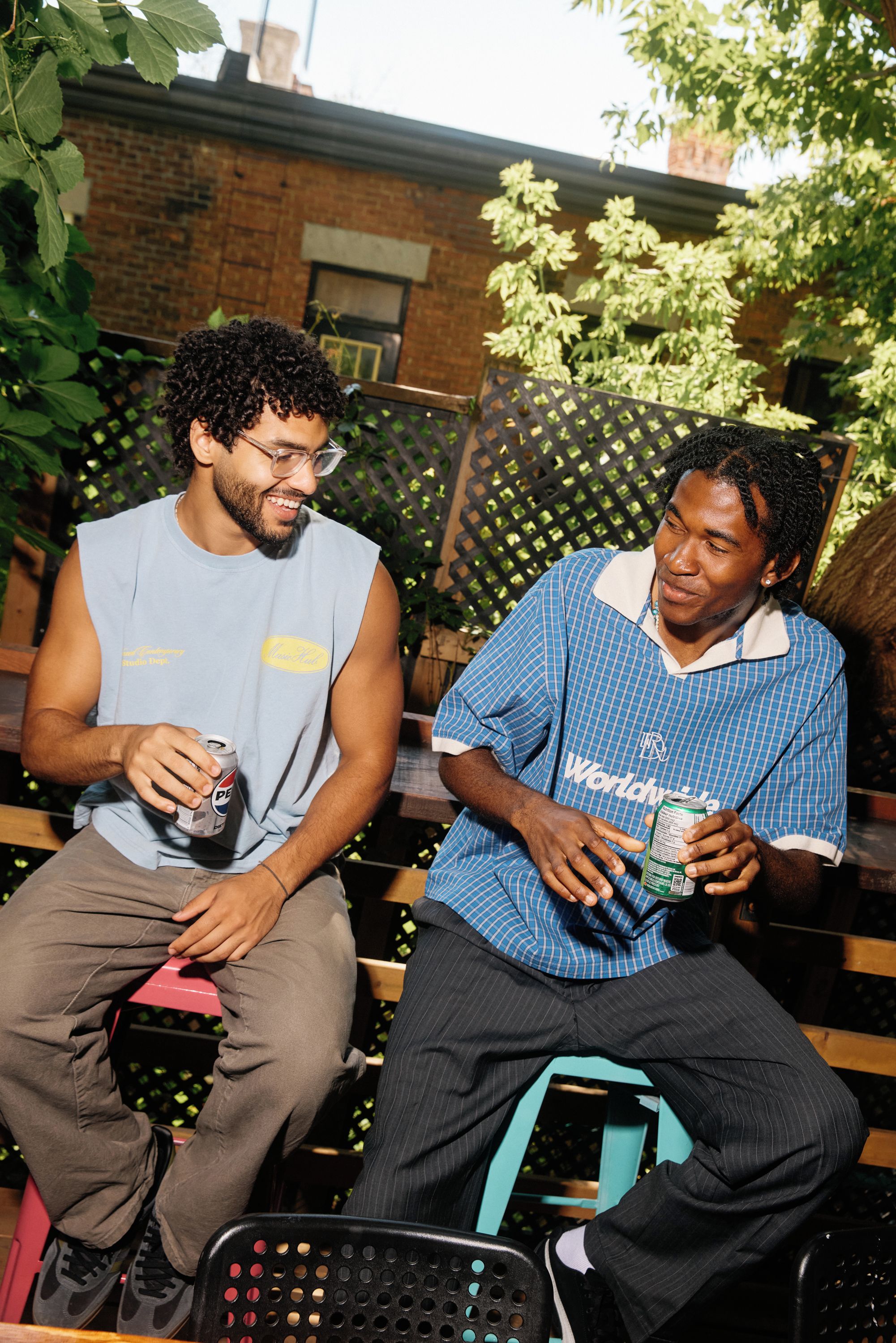 Two individuals seated on a bench outdoors, one holding a can and the other holding a carton.