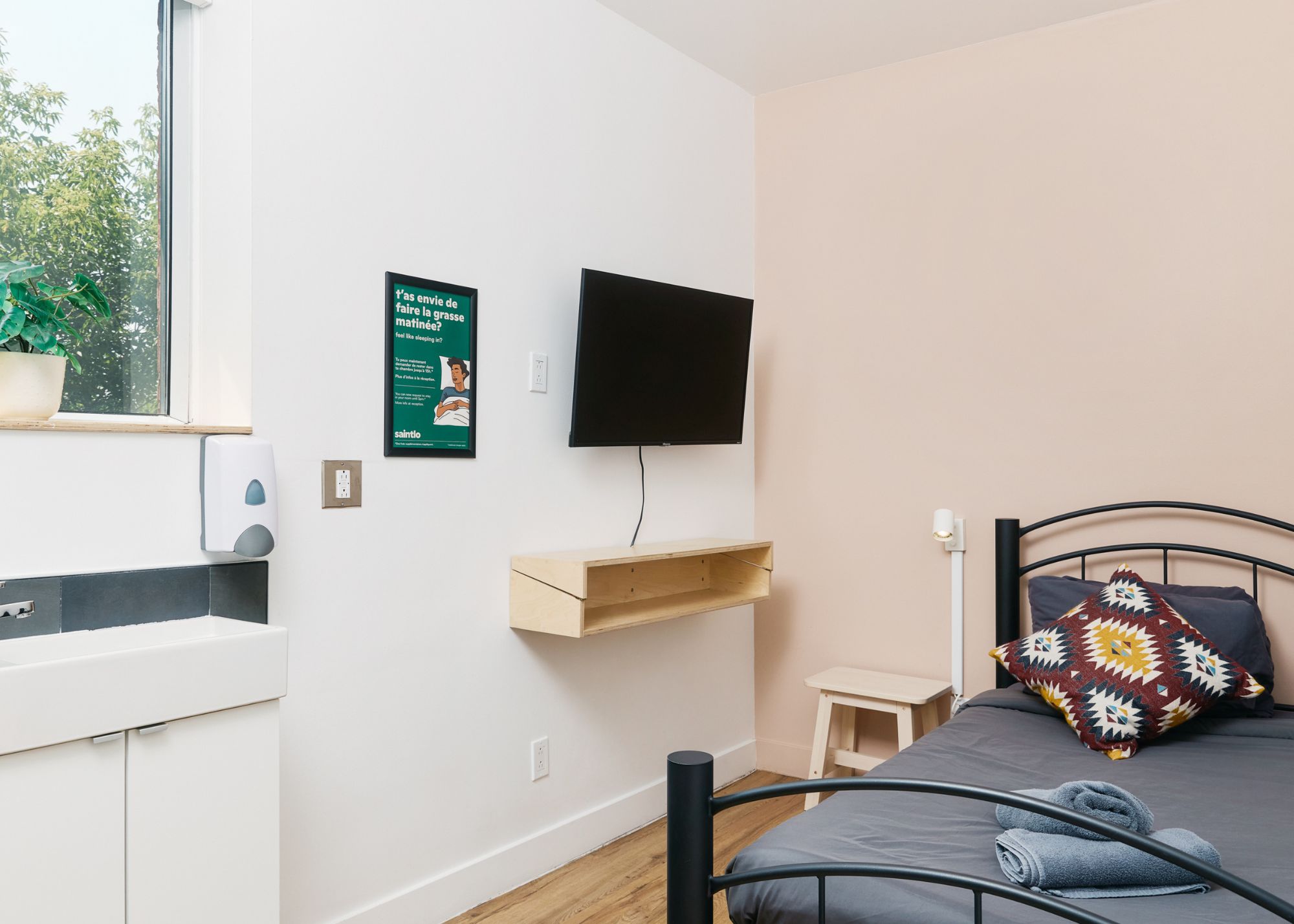 Una camera da letto con un letto, una televisione a muro, una piccola mensola in legno e una pianta in vaso sul davanzale.