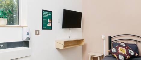 Una camera da letto con un letto, una televisione a muro, una piccola mensola in legno e una pianta in vaso sul davanzale.