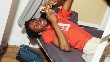A person lying on a bunk bed with a smartphone, a blue bag, and black slippers on the floor.