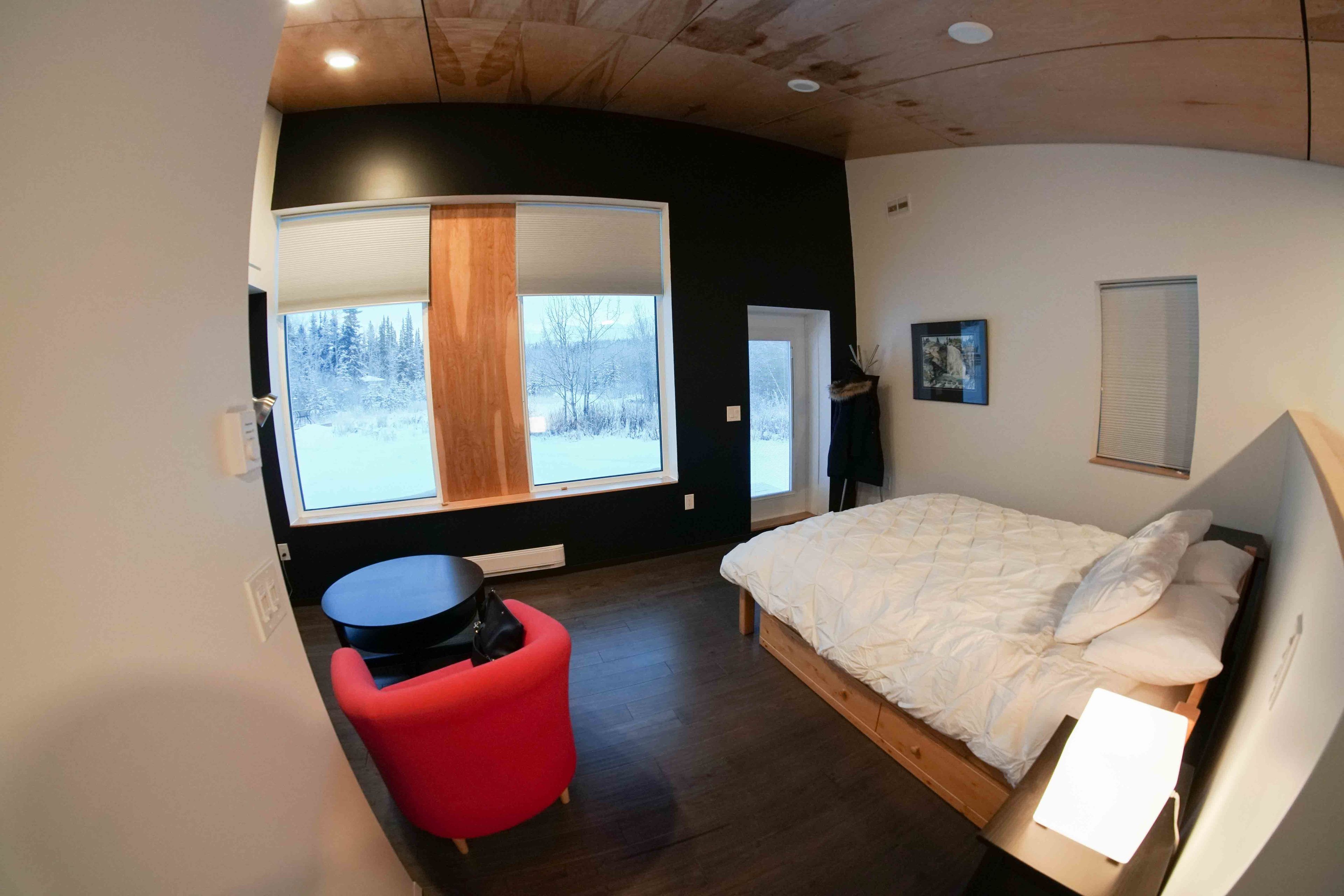 Ein modernes Schlafzimmer mit einem großen Bett, einem roten Sessel, einem kleinen Tisch und Blick auf eine verschneite Landschaft durch ein großes Fenster.