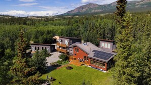 A modern multi-story house with a large deck, surrounded by a forested landscape and mountains in the background. - Boréale Ranch (Whitehorse)