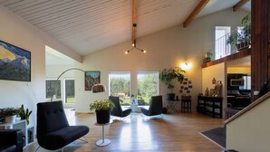 A spacious living room with a high ceiling, wooden flooring, and modern furniture. There are large windows providing natural light and views of the outdoors. - Boréale Ranch (Whitehorse)