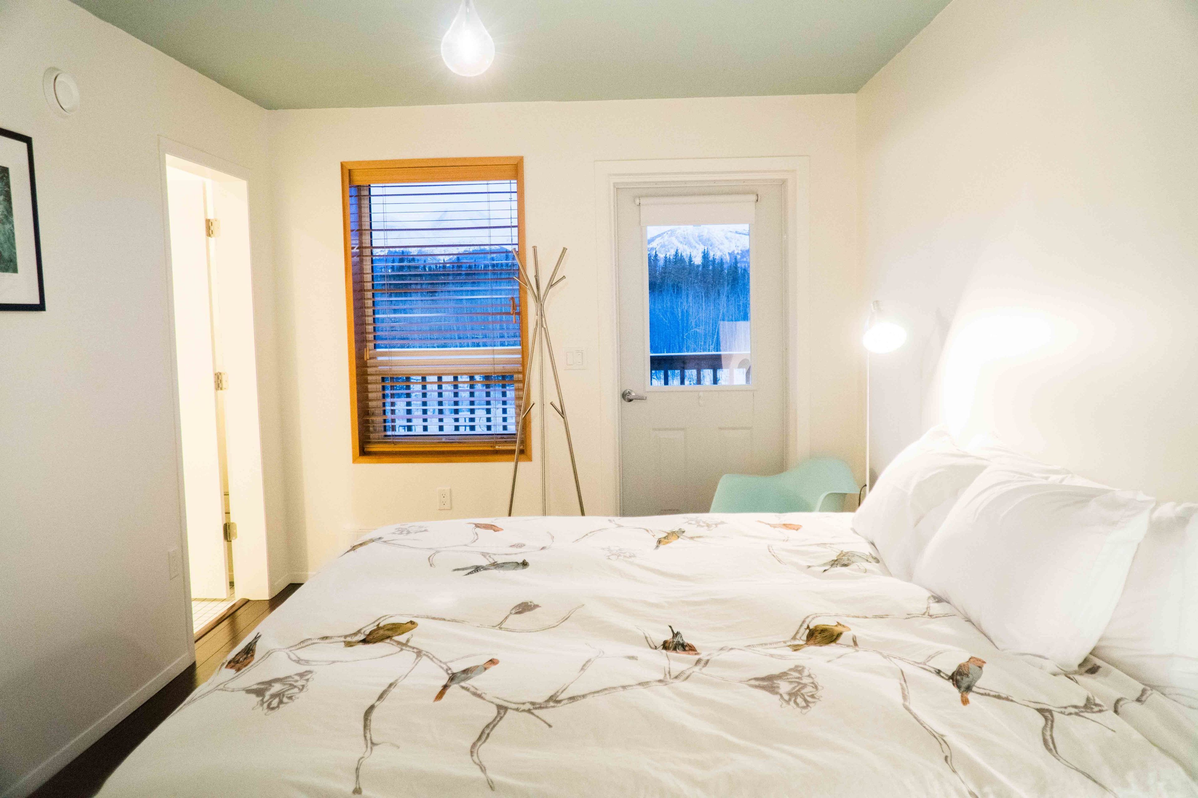 A bedroom with a bed, a window with blinds, a door, and a view of a snowy mountain.