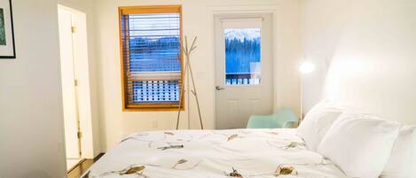 A bedroom with a bed, a window with blinds, a door, and a view of a snowy mountain.