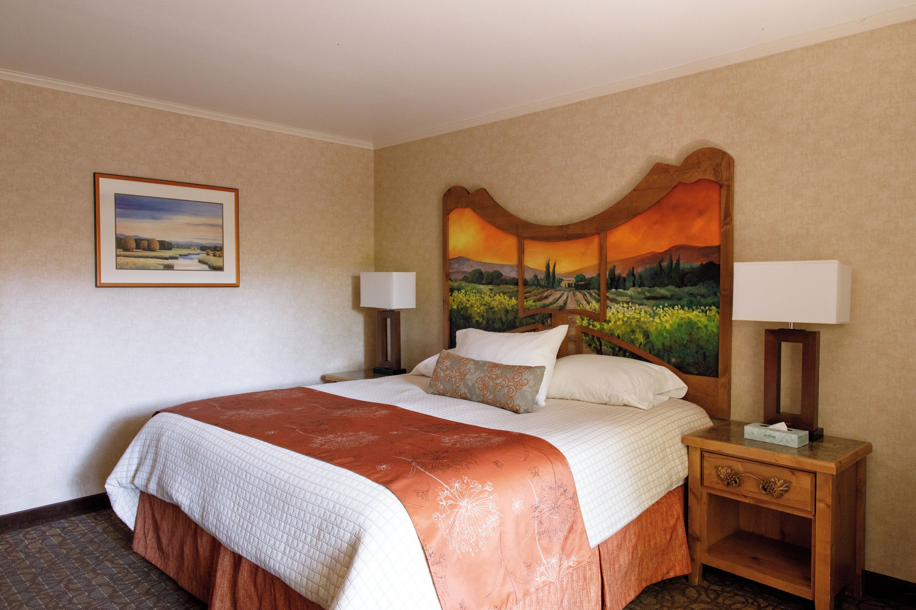 A hotel room with a large bed, a headboard with a sunset painting, two bedside lamps, and a framed landscape painting on the wall.