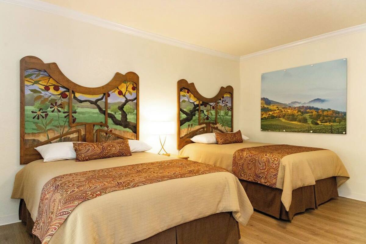 A hotel room with two beds, each with a patterned bedspread and matching pillows. There are two wall-mounted paintings depicting landscapes.