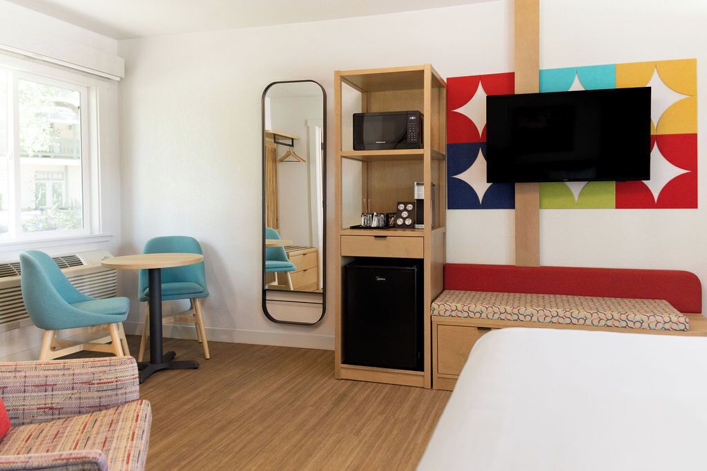 Une chambre d’hôtel équipée d’un lit, d’un petit bureau avec une chaise, d’un four à micro-ondes, d’un réfrigérateur, d’une télévision et d’une décoration murale colorée.