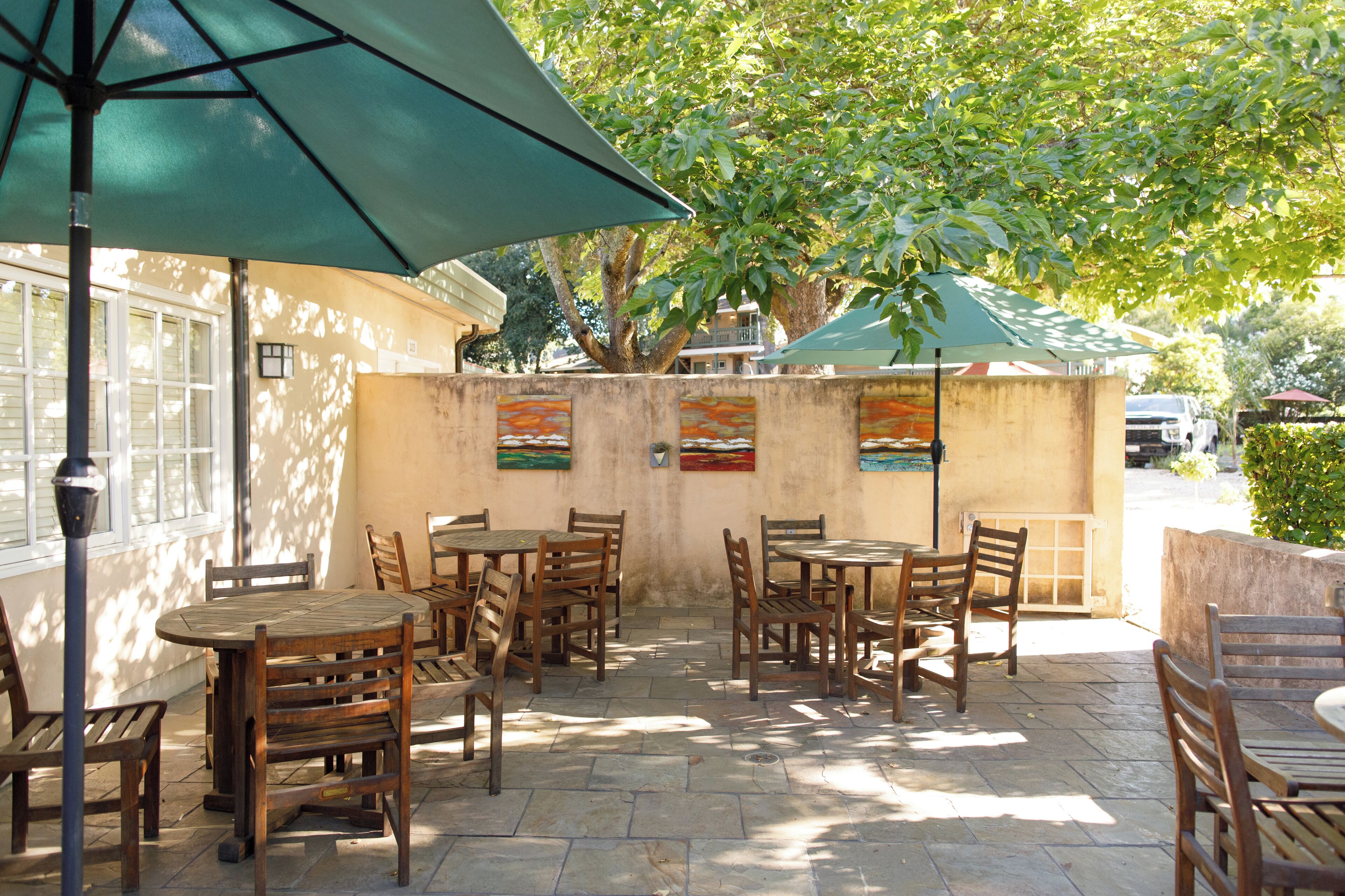 An outdoor patio with wooden tables and chairs, a green umbrella, and artwork on the wall.
