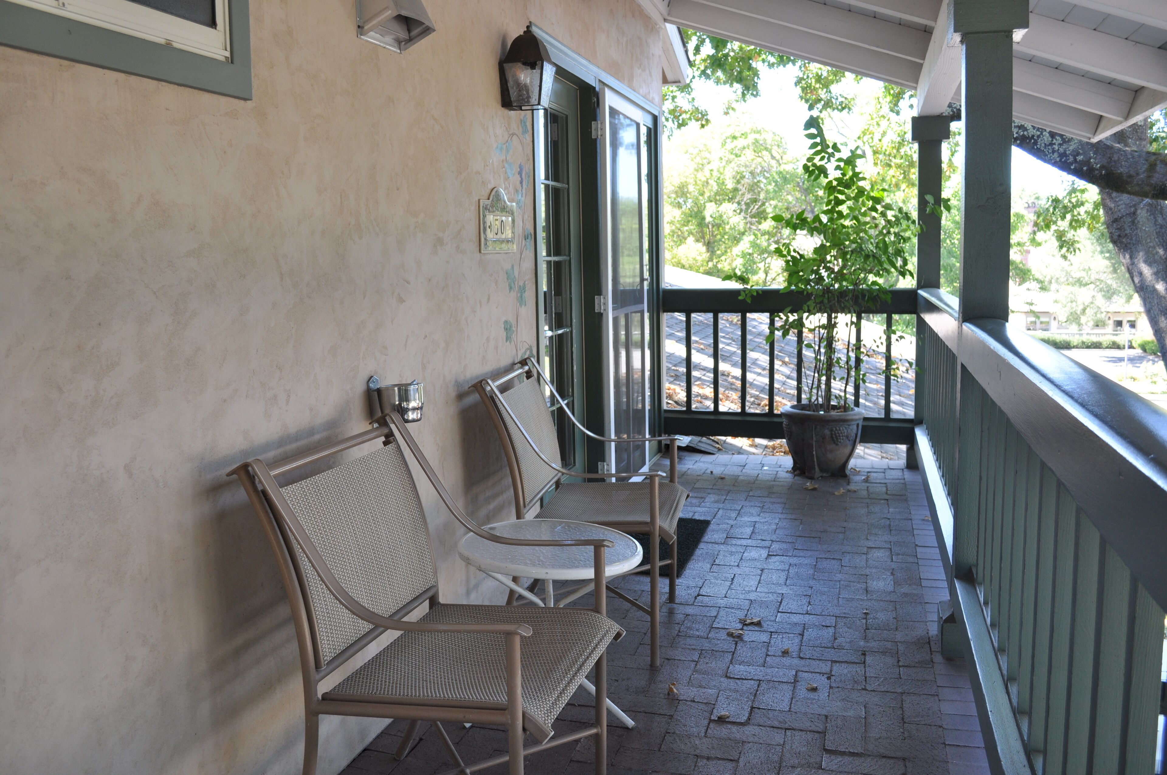 A covered outdoor seating area with metal chairs and a small table, a potted plant, and a view of trees and buildings in the background.