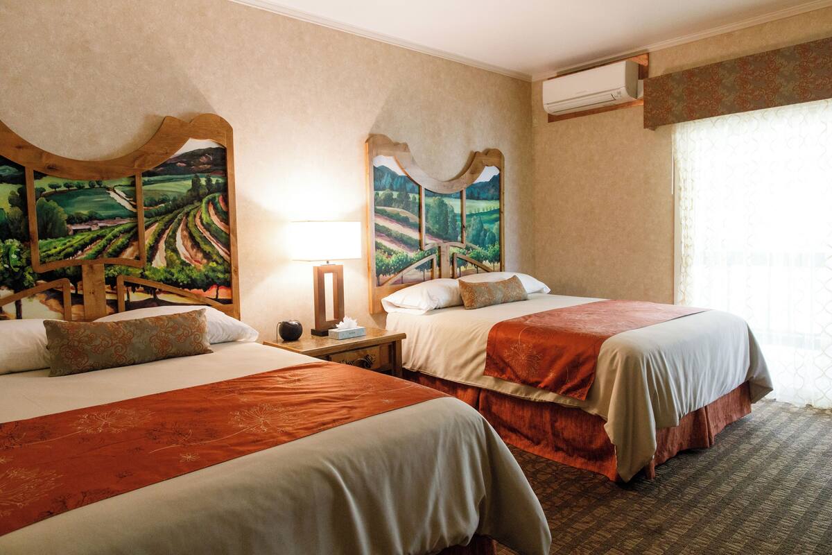 A hotel room with two beds, a nightstand, a lamp, and a large landscape painting on the wall.