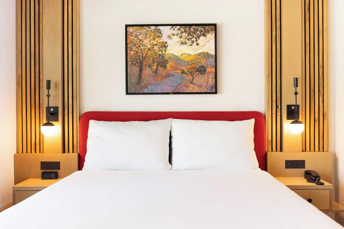 A hotel room with a large bed, a red headboard, two bedside tables with lamps, a painting on the wall, and a clock on the nightstand.