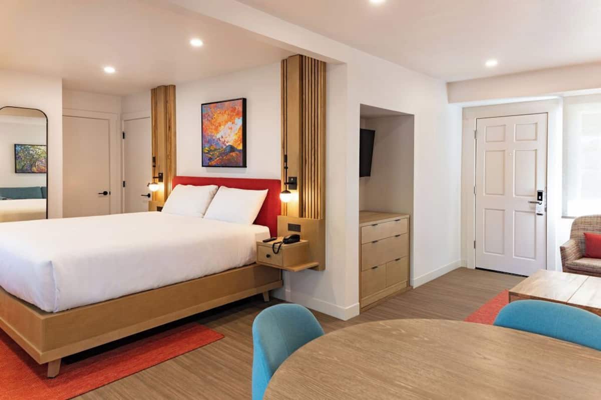 A modern hotel room with a large bed, bedside tables, a TV, and a dining area with chairs.